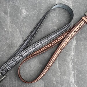 Op de afbeelding: Twee leren riemen met decoratieve stiksels en zilverkleurige gespen. E&eacute;n riem is grijs, de andere bruin. De riemen liggen op een grijze stenen ondergrond. De riemen hebben een klassieke western stijl.