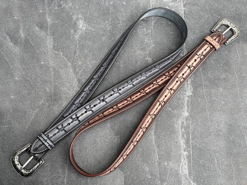 Op de afbeelding: Twee leren riemen met decoratieve stiksels en zilverkleurige gespen. E&eacute;n riem is grijs, de andere bruin. De riemen liggen op een grijze stenen ondergrond. De riemen hebben een klassieke western stijl.