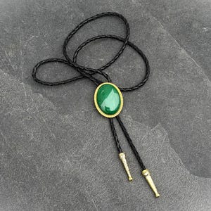 May include: A bolo tie featuring a large, oval, green stone set in a gold-colored frame. The tie has a black braided leather cord and gold-colored tips. The bolo tie is a classic accessory.