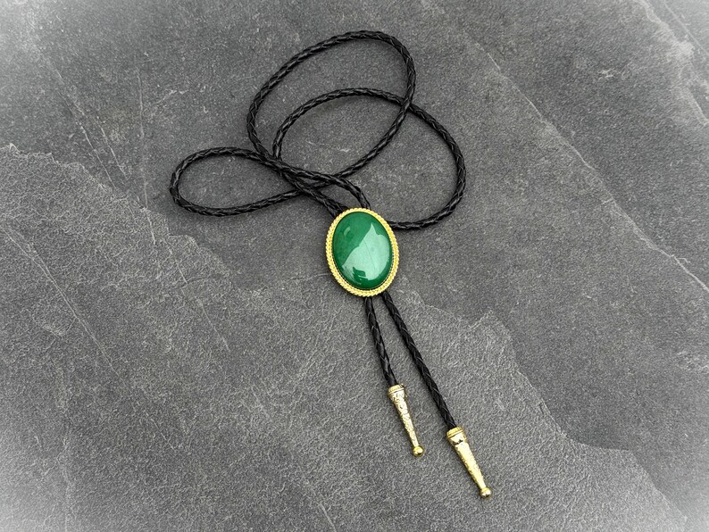 May include: A bolo tie featuring a large, oval, green stone set in a gold-colored frame. The tie has a black braided leather cord and gold-colored tips. The bolo tie is a classic accessory.