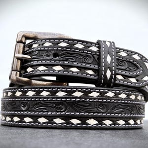 May include: A black leather belt with white stitching and a diamond pattern. The belt has an intricate floral design and a silver buckle.