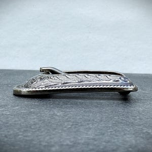 Puede incluir: Un clip de dinero de metal plateado con un dise&ntilde;o floral grabado y ornamentado. El clip tiene una forma curvada con una peque&ntilde;a palanca y un patr&oacute;n detallado, probablemente para guardar efectivo o tarjetas.