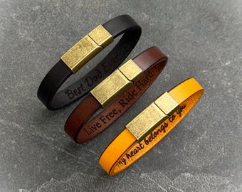 Puede incluir: Tres pulseras de cuero en negro, marrón y naranja, cada una con un cierre dorado. La pulsera negra dice "Best Dad Ever", la marrón "Live Free, Ride Hard" y la naranja "my heart belongs to you".