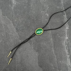 May include: A black bolo tie with a braided leather cord. The focal point is an oval, green stone set in a gold-colored frame. The tips of the cord are gold-colored.