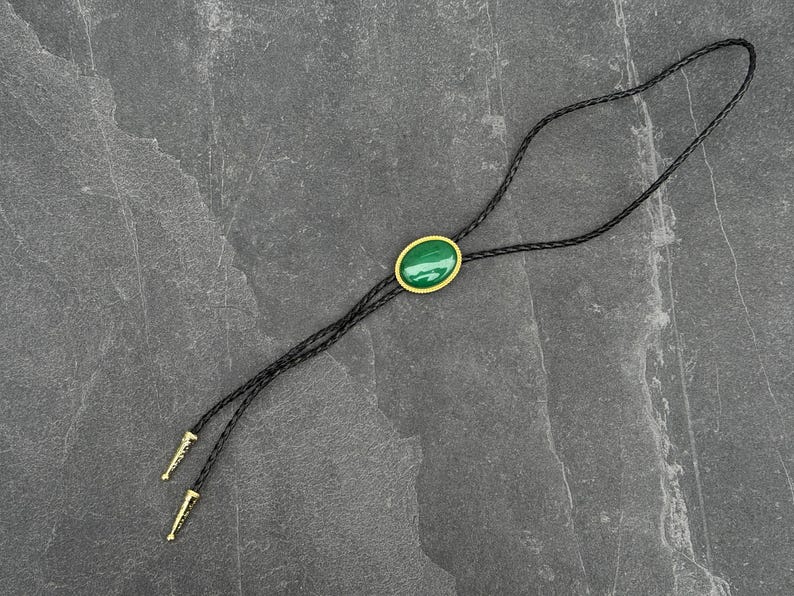 May include: A black bolo tie with a braided leather cord. The focal point is an oval, green stone set in a gold-colored frame. The tips of the cord are gold-colored.