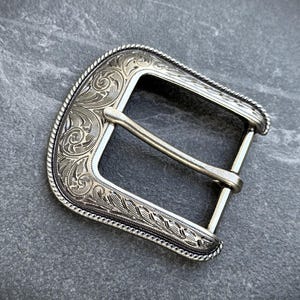 Puede incluir: Hebilla de cintur&oacute;n plateada con intrincado grabado floral y de volutas. La hebilla tiene forma rectangular con un borde curvo y una barra central para el cintur&oacute;n. El dise&ntilde;o incluye un borde en forma de cuerda.