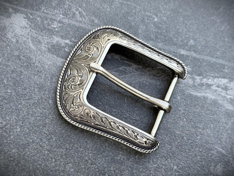 Puede incluir: Hebilla de cintur&oacute;n plateada con intrincado grabado floral y de volutas. La hebilla tiene forma rectangular con un borde curvo y una barra central para el cintur&oacute;n. El dise&ntilde;o incluye un borde en forma de cuerda.