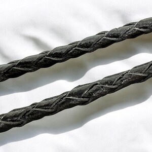 May include: Close-up of two black braided leather cords. The cords are thick and have a textured surface, with visible interwoven patterns. The background is a plain white surface, providing contrast to the dark cords.