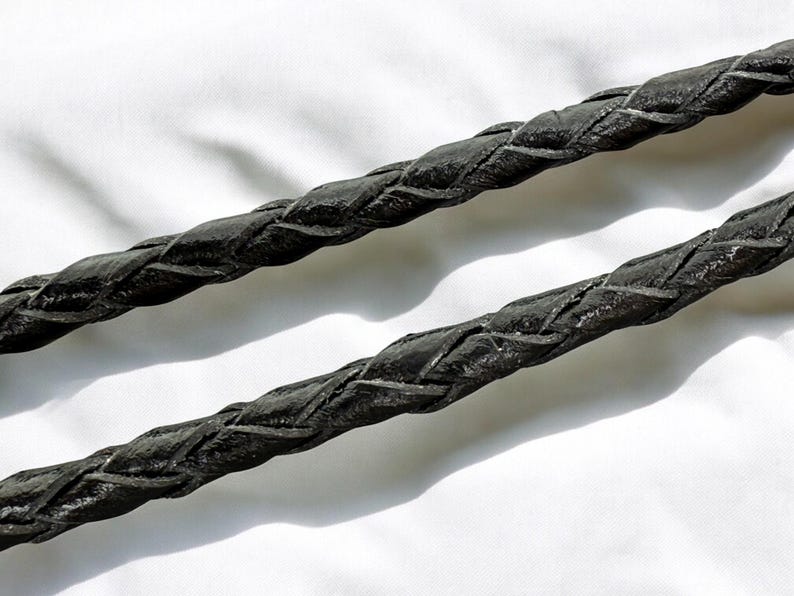 May include: Close-up of two black braided leather cords. The cords are thick and have a textured surface, with visible interwoven patterns. The background is a plain white surface, providing contrast to the dark cords.