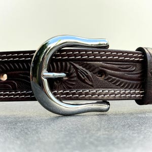 May include: A brown leather belt with a silver buckle. The belt has a floral pattern and white stitching.