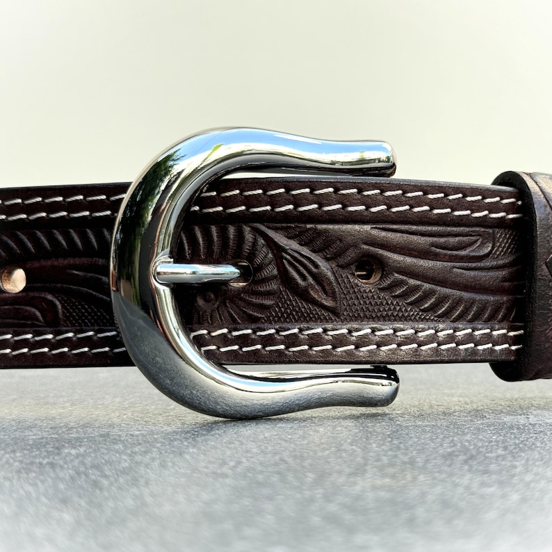 May include: A brown leather belt with a silver buckle. The belt has a floral pattern and white stitching.