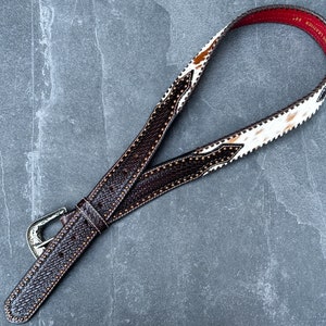 May include: A brown leather belt with a white and brown cowhide pattern. The belt has a silver buckle and is decorated with small metal studs.