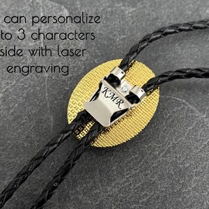 May include: A close-up of a bolo tie with a silver-colored clasp engraved with "KMR". The tie features a gold-colored oval pendant and black braided cords. The text in the image says, "You can personalize up to 3 characters inside with laser engraving."
