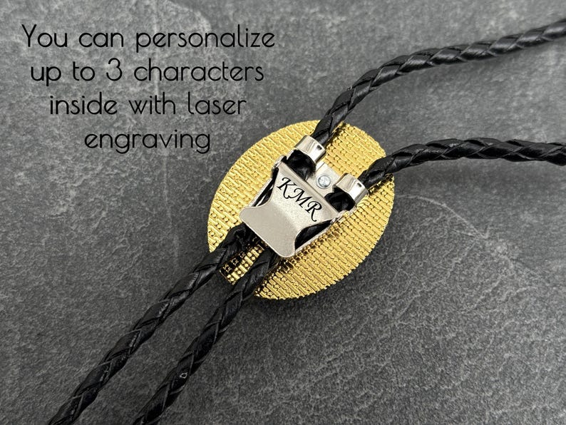 May include: A close-up of a bolo tie with a silver-colored clasp engraved with "KMR". The tie features a gold-colored oval pendant and black braided cords. The text in the image says, "You can personalize up to 3 characters inside with laser engraving."