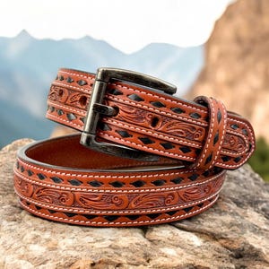 May include: A brown leather belt with a black stitched design and intricate floral tooling. The belt has a silver buckle and is folded in half.