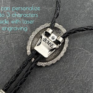 May include: A silver bolo tie with a braided black leather cord. The silver pendant has an oval shape with a decorative border and the initials "BME" engraved on the front. Text in the image says, "You can personalize up to 3 characters inside with laser engraving."