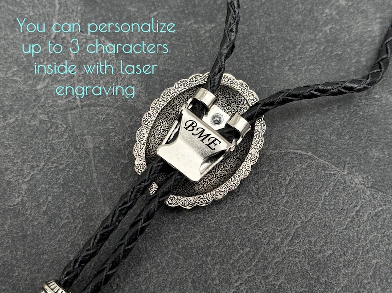 May include: A silver bolo tie with a braided black leather cord. The silver pendant has an oval shape with a decorative border and the initials "BME" engraved on the front. Text in the image says, "You can personalize up to 3 characters inside with laser engraving."