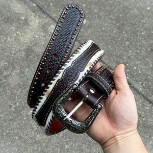 May include: A brown leather belt with a white and brown braided design and a silver buckle. The belt has small brass studs along the edge.