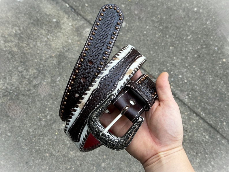 May include: A brown leather belt with a white and brown braided design and a silver buckle. The belt has small brass studs along the edge.