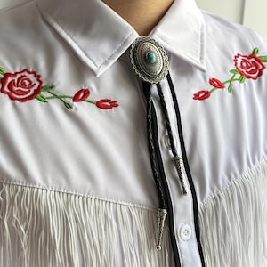 May include: White shirt with red rose embroidery and fringe detail. Features a silver bolo tie with a turquoise stone and black leather cord. The shirt has a classic collar and button-down front.