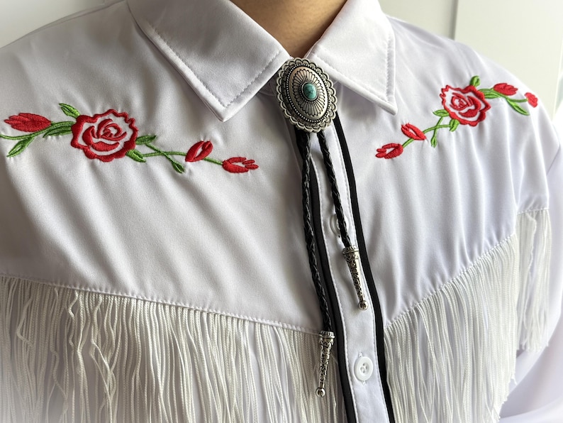 May include: White shirt with red rose embroidery and fringe detail. Features a silver bolo tie with a turquoise stone and black leather cord. The shirt has a classic collar and button-down front.