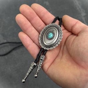 May include: Oval silver bolo tie with a turquoise stone center. The bolo tie has a decorative silver border and black braided cord. The tips of the bolo tie are silver with a decorative pattern.