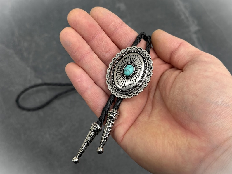 May include: Oval silver bolo tie with a turquoise stone center. The bolo tie has a decorative silver border and black braided cord. The tips of the bolo tie are silver with a decorative pattern.