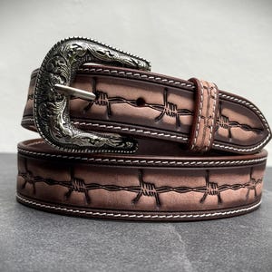 Op de afbeelding: Een bruine leren riem met een zilveren gesp. De riem heeft een prikkeldraadontwerp en witte stiksels. De gesp heeft een sierlijk, gegraveerd patroon. Een accessoire in westernstijl.