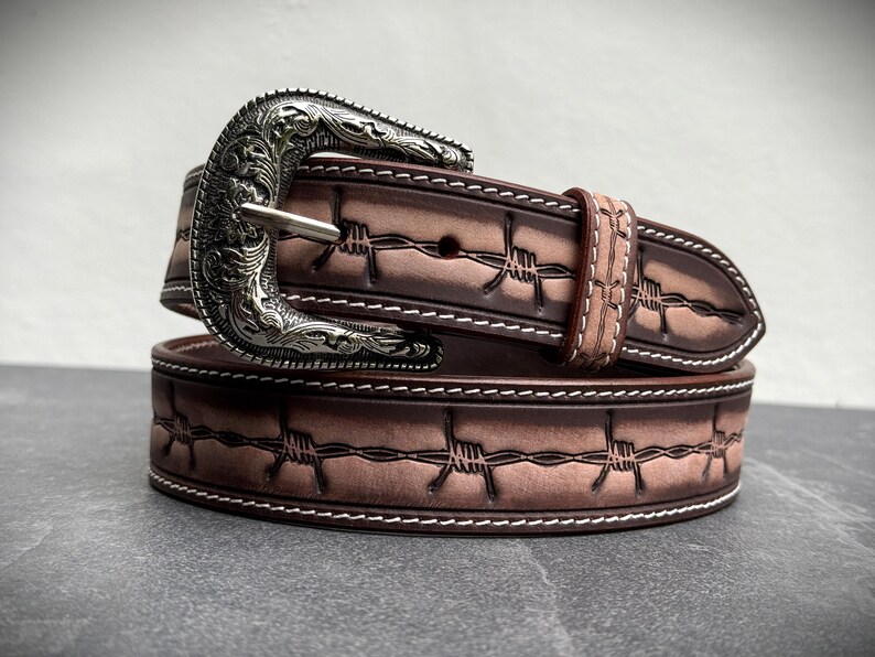 Op de afbeelding: Een bruine leren riem met een zilveren gesp. De riem heeft een prikkeldraadontwerp en witte stiksels. De gesp heeft een sierlijk, gegraveerd patroon. Een accessoire in westernstijl.