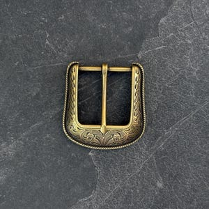 Puede incluir: Hebilla de cintur&oacute;n decorativa de color lat&oacute;n con un dise&ntilde;o floral ornamentado. La hebilla tiene forma rectangular con una barra central y un borde detallado. El metal tiene un acabado vintage y antiguo.