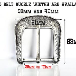 Puede incluir: Hebilla de cintur&oacute;n plateada con un dise&ntilde;o ornamentado. La hebilla tiene forma rectangular con esquinas redondeadas y una barra central. El texto indica dos anchos disponibles: 38 mm o 42 mm. La hebilla mide 61 mm de ancho y 63 mm de alto.