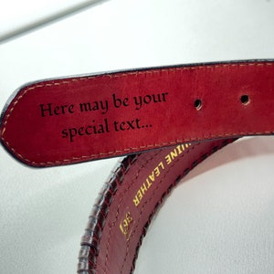 May include: A close-up of a brown leather belt with a red underside. The belt has a braided design and the words "Genuine Leather" are embossed on the side. The underside of the belt has the text "Here may be your special text..." engraved on it.