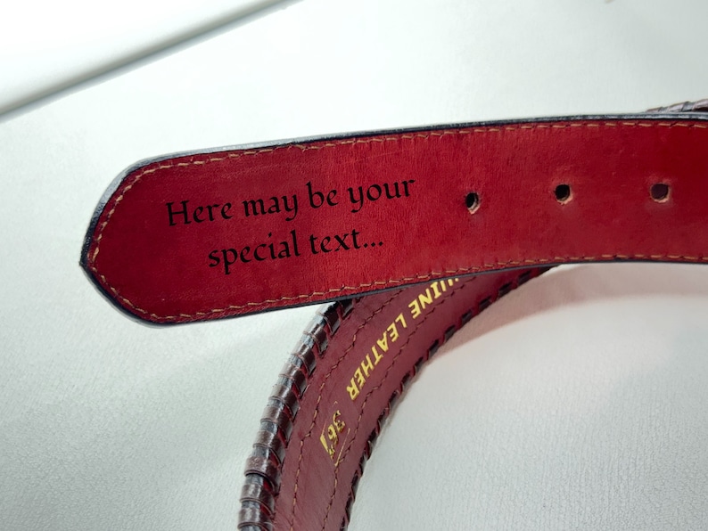 May include: A close-up of a brown leather belt with a red underside. The belt has a braided design and the words "Genuine Leather" are embossed on the side. The underside of the belt has the text "Here may be your special text..." engraved on it.