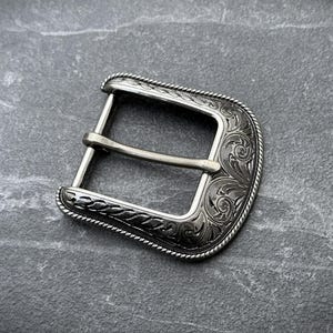 Puede incluir: Hebilla de cintur&oacute;n plateada con un dise&ntilde;o grabado y ornamentado. La hebilla tiene forma rectangular con bordes redondeados y una barra central para el cintur&oacute;n. Los bordes est&aacute;n detallados con un patr&oacute;n similar a una cuerda.