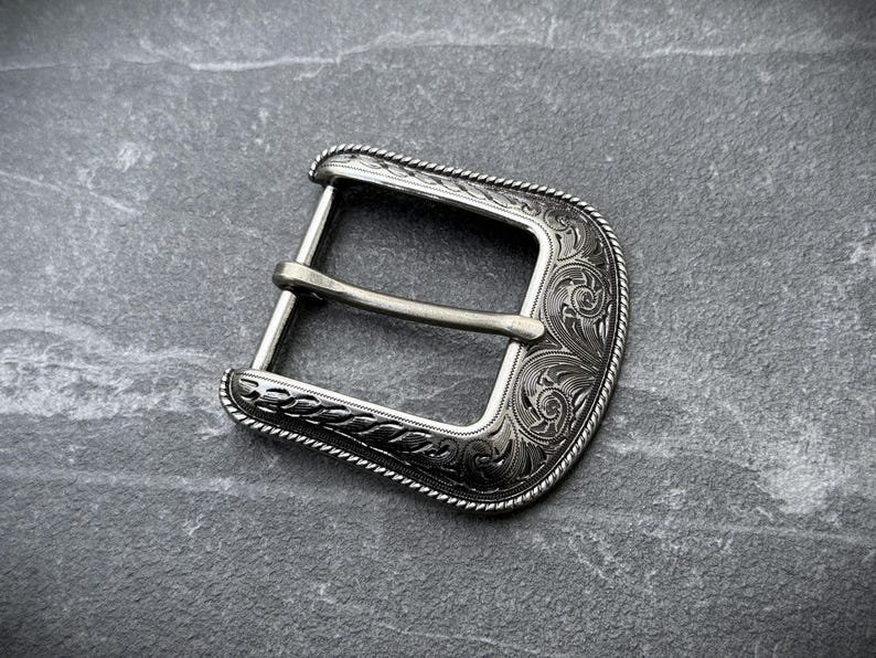 Puede incluir: Hebilla de cintur&oacute;n plateada con un dise&ntilde;o grabado y ornamentado. La hebilla tiene forma rectangular con bordes redondeados y una barra central para el cintur&oacute;n. Los bordes est&aacute;n detallados con un patr&oacute;n similar a una cuerda.