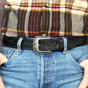 Puede incluir: Un cintur&oacute;n de cuero negro con una hebilla plateada con un dise&ntilde;o floral ornamentado. El cintur&oacute;n se usa con vaqueros azules y una camisa a cuadros. El cintur&oacute;n tiene un patr&oacute;n en relieve detallado.