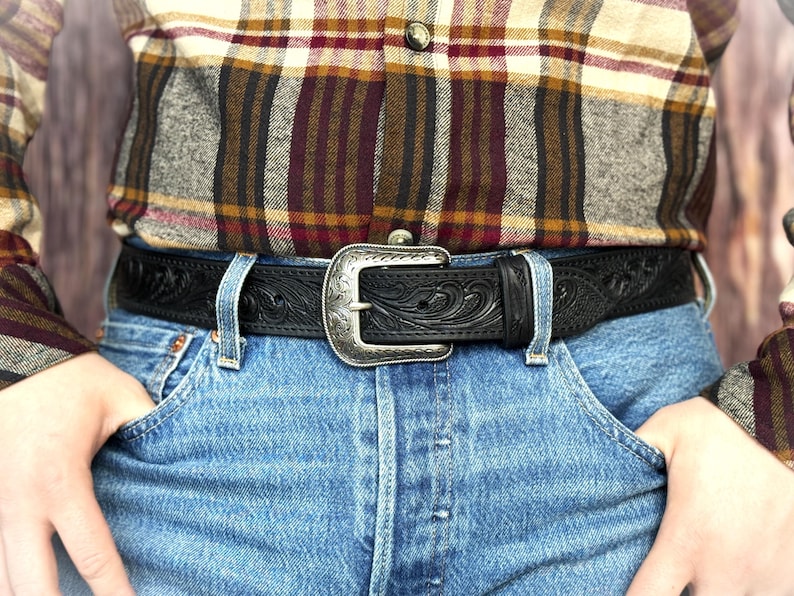 Puede incluir: Un cintur&oacute;n de cuero negro con una hebilla plateada con un dise&ntilde;o floral ornamentado. El cintur&oacute;n se usa con vaqueros azules y una camisa a cuadros. El cintur&oacute;n tiene un patr&oacute;n en relieve detallado.