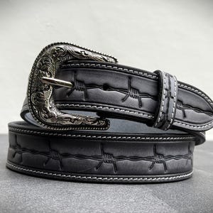 Op de afbeelding: Een grijze leren riem met een zilverkleurige gesp. De riem heeft een prikkeldraadontwerp en witte stiksels. De gesp heeft een sierlijk, gegraveerd patroon. De riem is opgerold en toont het ontwerp.