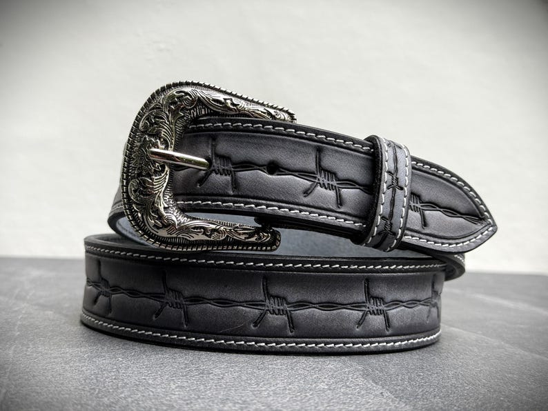 Op de afbeelding: Een grijze leren riem met een zilverkleurige gesp. De riem heeft een prikkeldraadontwerp en witte stiksels. De gesp heeft een sierlijk, gegraveerd patroon. De riem is opgerold en toont het ontwerp.