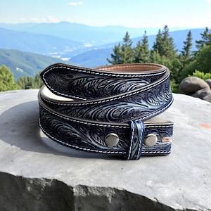 May include: A dark blue leather belt featuring intricate floral tooling and embossed patterns. The belt is finished with white contrast stitching along the edges and includes two silver-toned metal snaps on the keeper loop for secure fastening.