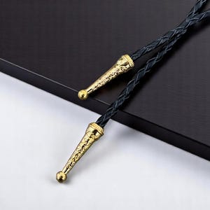 May include: A black bolo tie with gold-colored metal tips. The tips have an ornate design and a small, round ball at the end. The tie itself is made of braided black leather.