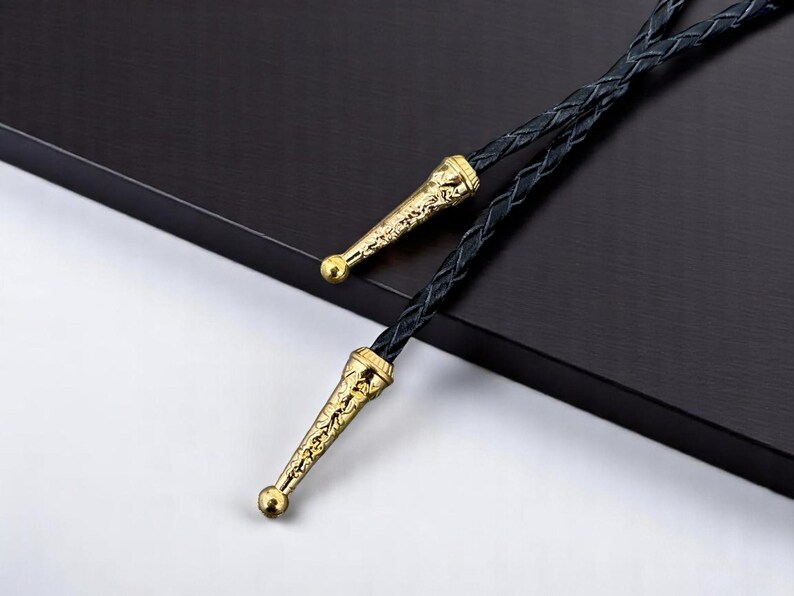 May include: A black bolo tie with gold-colored metal tips. The tips have an ornate design and a small, round ball at the end. The tie itself is made of braided black leather.