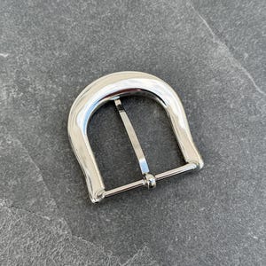 May include: A silver metal belt buckle with a smooth, rounded design. The buckle has a single prong and a simple, functional style.