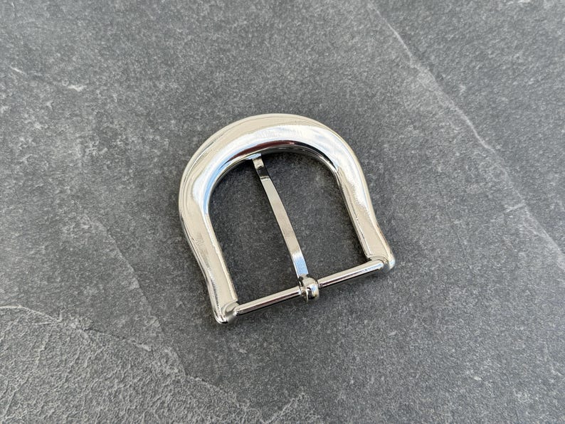 May include: A silver metal belt buckle with a smooth, rounded design. The buckle has a single prong and a simple, functional style.