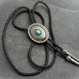 Op de afbeelding: Een zilveren bolo stropdas met een ovale turquoise steen in het midden. De zilveren hanger heeft een decoratieve geschulpte rand. Het zwarte gevlochten leren koord heeft zilveren uiteinden.