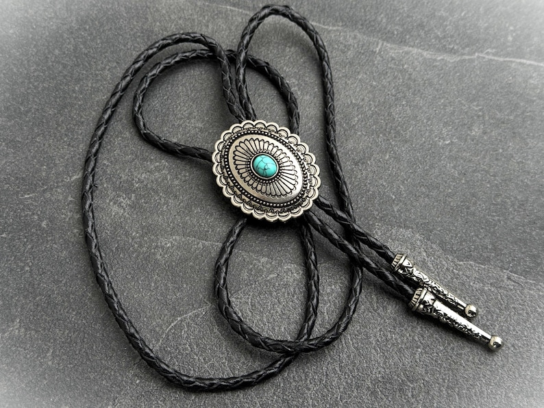 May include: A silver bolo tie with an oval turquoise stone center. The silver pendant has a decorative scalloped edge. The black braided leather cord has silver tips.