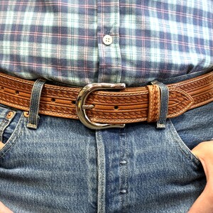 May include: A brown leather belt with a silver buckle and a woven design. The belt is worn over a pair of blue jeans.