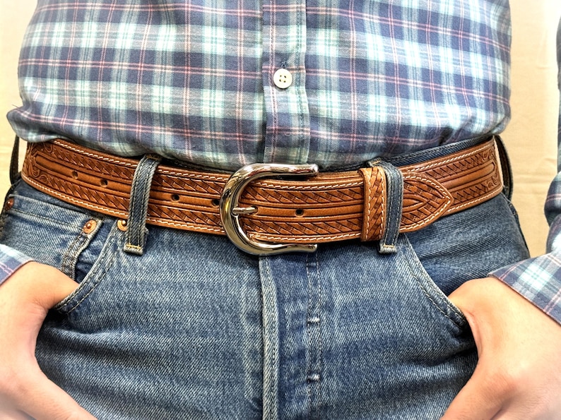 May include: A brown leather belt with a silver buckle and a woven design. The belt is worn over a pair of blue jeans.