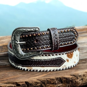 May include: A brown leather belt with a silver buckle and a white and brown cowhide pattern. The belt has a red stitching detail and is decorated with small metal studs.