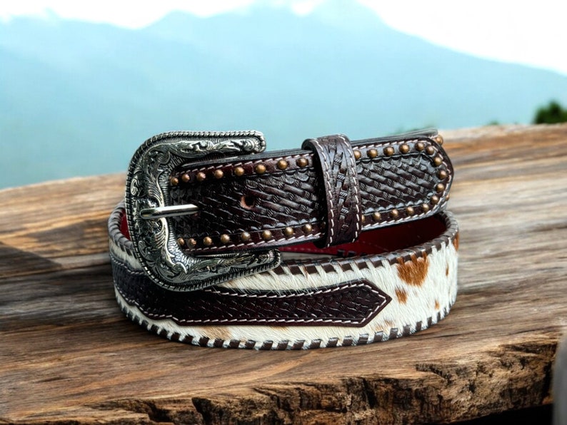 May include: A brown leather belt with a silver buckle and a white and brown cowhide pattern. The belt has a red stitching detail and is decorated with small metal studs.